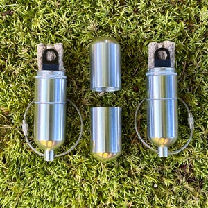 May include: Two cylindrical silver metal fire starters with exposed wicks and attached wire key rings, positioned alongside a separate silver metal cap and base component. The items feature a smooth, brushed metallic finish and a compact design.