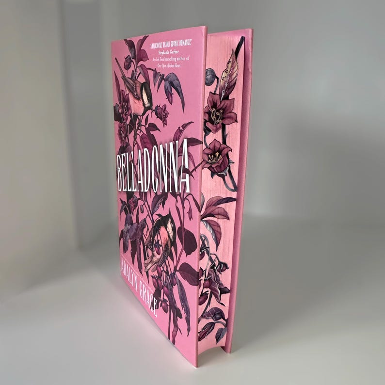 May include: A pink hardcover book with the title "BELLADONNA" in white lettering. The cover features a floral design with pink flowers and birds.