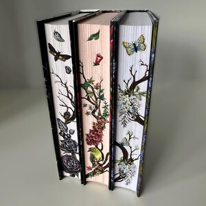 May include: Three hardcover books with floral and insect designs on the spines. The spines are white, pink, and blue with black trim. The designs include butterflies, birds, and flowers.