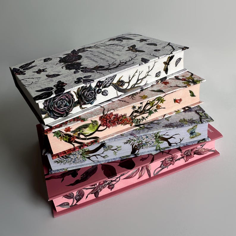 May include: A stack of five hardcover journals with floral and botanical designs. The top journal has a black and white design with a quote about nature. The second journal has a pink and white design with a bird and flowers. The third journal has a blue and white design with flowers and leaves. The fourth journal has a pink and white design with flowers and leaves. The bottom journal has a pink and white design with flowers and leaves.