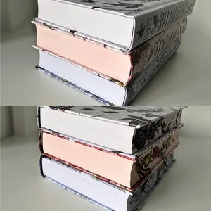 May include: Three hardcover books stacked on top of each other. The top book has a white cover with black floral designs and the text "A Ballad of Love and Glory" printed on the spine. The middle book has a pink cover with black floral designs and the text "The Hollow" printed on the spine. The bottom book has a blue cover with black floral designs and the text "The Raven Cycle" printed on the spine.