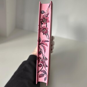 May include: The spine of a pink book with a floral design. The design features pink flowers and leaves on a pink background.