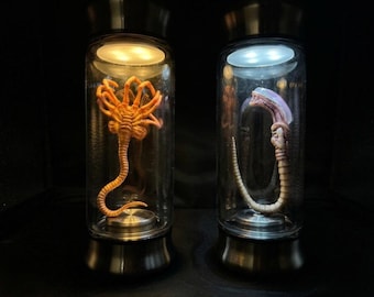 May include: Two cylindrical glass display canisters with metallic bases and lids. The left canister contains an orange, multi-legged creature illuminated by warm light. The right canister holds a pale, curved creature lit by cool white light.