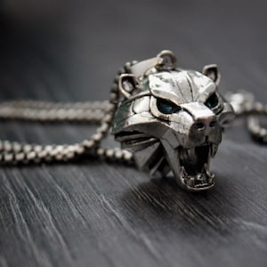 May include: A silver necklace with a wolf head pendant. The pendant has blue eyes and an open mouth with sharp teeth.