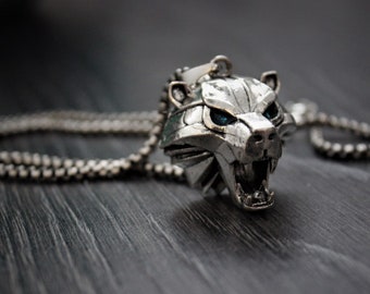 May include: A silver necklace with a wolf head pendant. The pendant has blue eyes and an open mouth with sharp teeth.
