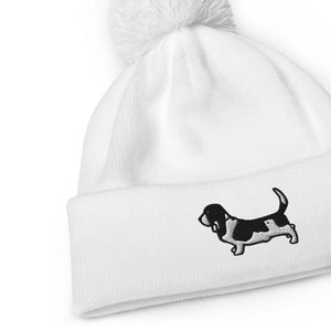 Könnte beinhalten: Weiße Strickmütze mit einem schwarzen und weißen gestickten Basset Hound auf der Vorderseite und einem weißen Bommel oben.