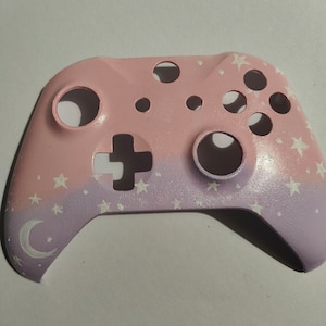 May include: A pink and purple video game controller with a white crescent moon and stars painted on it.