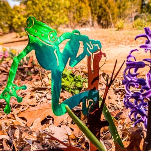 Puede incluir: Una escultura de rana de metal con un acabado verde y azul está posada en una rama de árbol de metal. La escultura está colocada en un jardín con hojas marrones y una flor de jacinto púrpura.