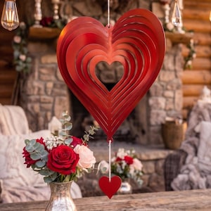 May include: A red metal heart-shaped wind spinner featuring concentric heart cutouts, suspended by a white string. A smaller, solid red heart hangs from the bottom of the larger structure, creating a layered, symmetrical design.