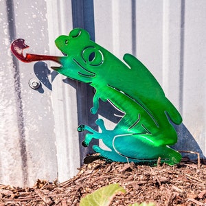 Puede incluir: Una escultura de rana de metal verde con una larga lengua que sobresale, sentada sobre un lecho de virutas de madera marrón. La rana está colocada contra una pared de metal plateado.