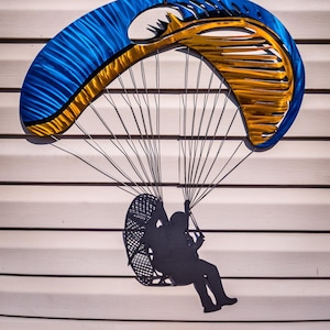 May include: A metal wall sculpture of a paraglider with a blue and gold canopy and a black silhouette of a person in a harness. The sculpture is mounted on a white wall.