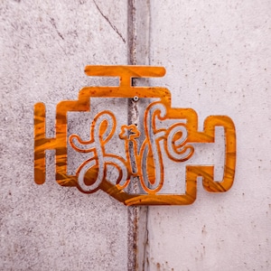 Pode incluir: Um recorte de metal de uma luz de controle do motor de um carro com a palavra "life" escrita dentro. O recorte é laranja e está montado em uma parede branca.