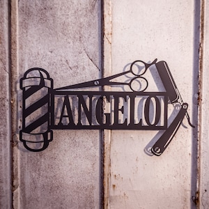 Puede incluir: Un letrero de metal negro con la palabra "ANGELOT" y un diseño de poste de barbero y tijeras.
