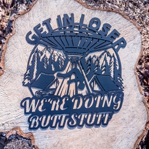 May include: Black metal sign with a UFO, trees, and the text "Get in loser, we're doing butt stuff".