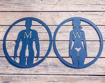May include: Two black metal signs with silhouettes of people in circles. The sign on the left has the letter "M" in the center of the silhouette. The sign on the right has the letter "V" in the center of the silhouette.