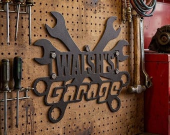 May include: A black metal wall sign featuring two crossed wrenches and the text "WALSH'S Garage" mounted on a brown pegboard. The sign is positioned among various hand tools, a red metal tool chest, and scattered hardware on a wooden workbench.