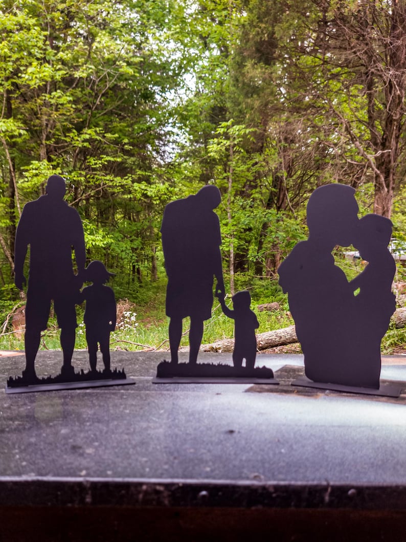 May include: Three black metal silhouettes of families. The first silhouette shows a father and son holding hands. The second silhouette shows a father and son holding hands. The third silhouette shows a father and child embracing.