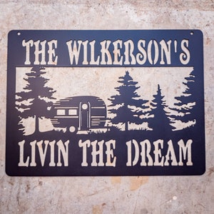 May include: Black metal sign with the text "The Wilkerson's Livin the Dream" in white. The sign features a silhouette of a camper and trees.