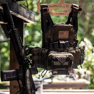 Könnte beinhalten: Eine beige taktische Weste mit einem US-Flaggen-Patch, die an einem Metallbügel mit dem aufgedruckten Wort "RIGHTNIT" hängt. Ein schwarzes Gewehr mit einem beigefarbenen Riemen lehnt an einer Holzoberfläche.