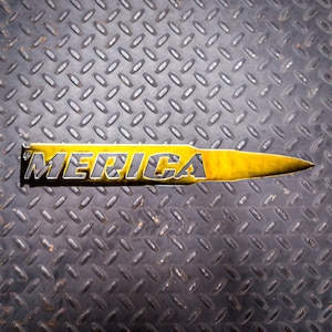 May include: A yellow metal bullet with the word "MERICA" cut out of it. The bullet is on a gray textured surface.