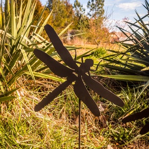 May include: Black metal dragonfly garden stake silhouette. The stake is placed in the ground with green grass and brown leaves around it.