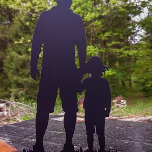 Pu&ograve; includere: Silhouette in metallo nero di un padre e un figlio in piedi uno accanto all'altro. La silhouette &egrave; posizionata su uno sfondo sfocato di alberi verdi.
