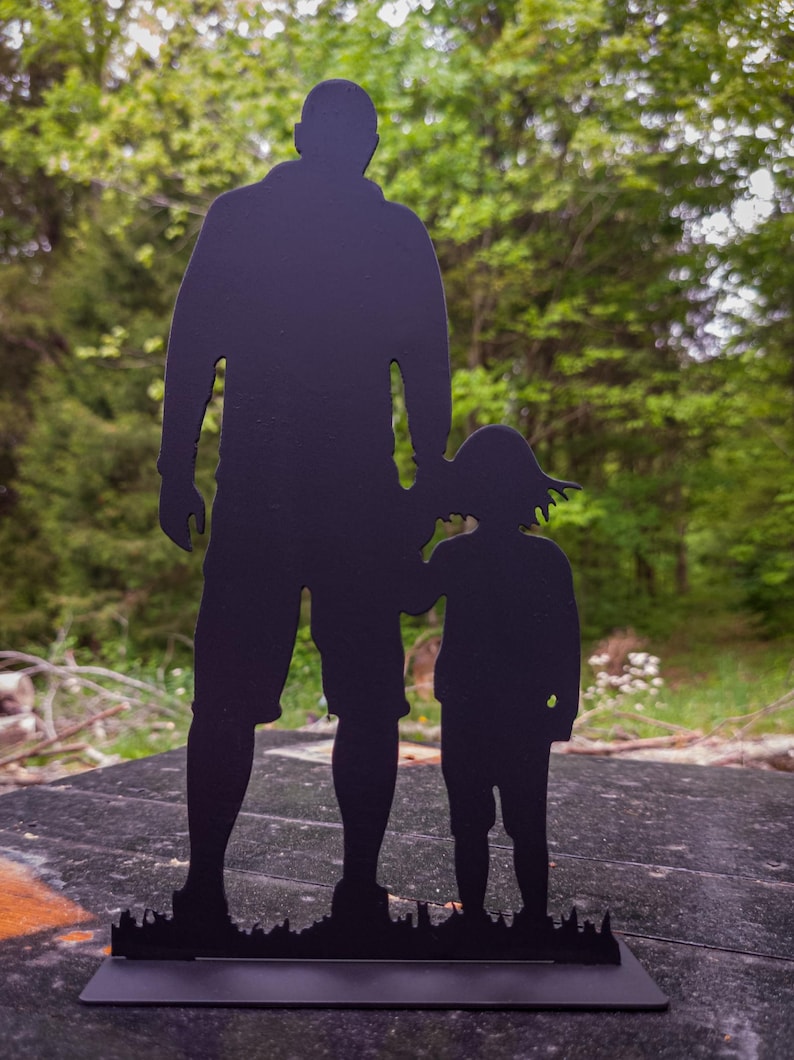 Pu&ograve; includere: Silhouette in metallo nero di un padre e un figlio in piedi uno accanto all'altro. La silhouette &egrave; posizionata su uno sfondo sfocato di alberi verdi.