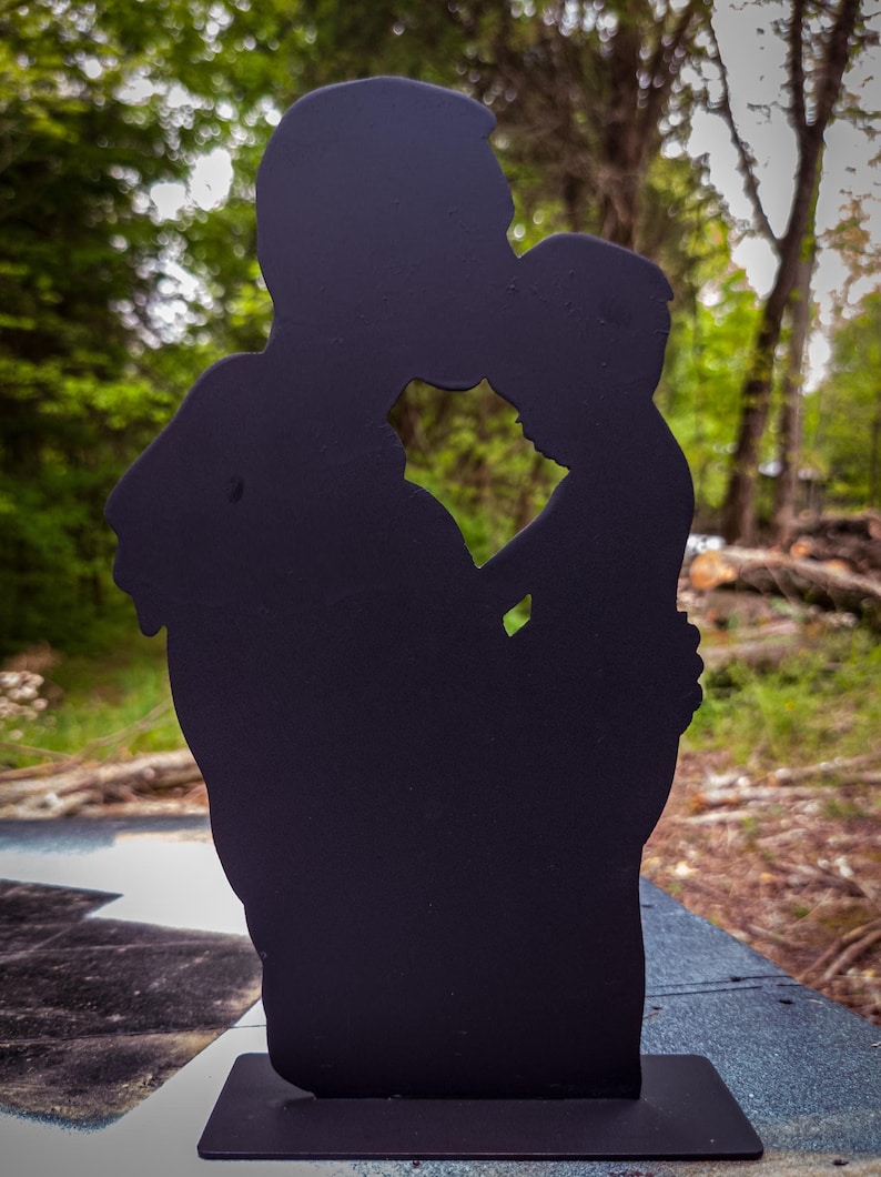 Pu&ograve; includere: Silhouette in metallo nero di un genitore che tiene un bambino. La silhouette &egrave; ritagliata a forma di genitore e figlio che si abbracciano.