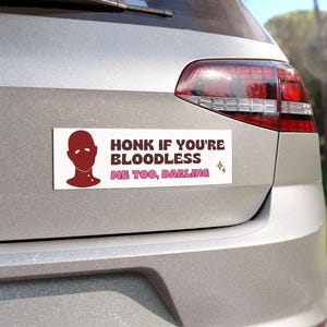 May include: A white bumper sticker with a red silhouette of a person's head with a pink mask. The text on the sticker reads "Honk if you're bloodless Me too, darling" with two yellow stars.