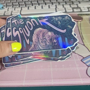 May include: A stack of iridescent stickers featuring a cartoon creature with the text "The Squonk" and an arrow pointing to the right.