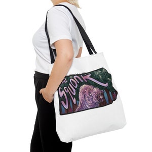 May include: A white tote bag with black straps featuring a colorful illustration of a pink creature with the text "The Squonk" in a pink and purple font.