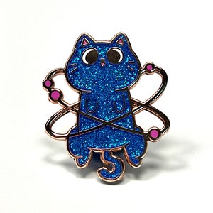May include: A blue glitter cat with pink accents is depicted as an atom. The cat has large eyes and a small tail. The atom is represented by a circular shape with pink dots around it.