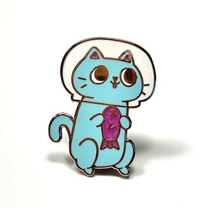 May include: A blue cat wearing a white astronaut helmet holds a pink glittery fish. The cat has large brown eyes and a pink tongue sticking out. The pin is made of metal with a gold finish.