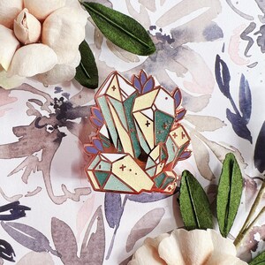 May include: A gold-toned enamel pin featuring a cluster of green, blue, and yellow geometric crystals with a glittery finish. The crystals are surrounded by small purple leaves.