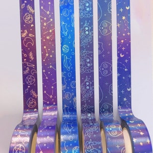 May include: Six rolls of iridescent holographic washi tape with different space-themed designs. The tape is blue, purple, and pink with white designs of stars, planets, cats, and skulls.