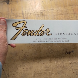 May include: A white Fender Stratocaster guitar decal with gold lettering. The decal reads "Fender Stratocaster" with "WITH SYNCHRONIZED TREMOLO" and "PAT. 2,573,254 2,741,146 2,960,900 3,143,028" underneath.