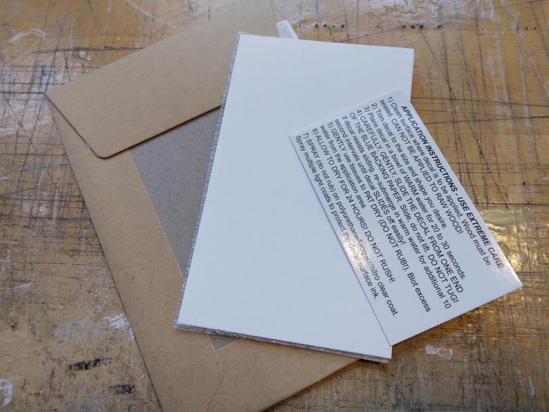 May include: Close-up of a white card with black text outlining application instructions for a decal. The text reads: "APPLICATION INSTRUCTIONS USE EXTREME CARE 1) Clean surface where decal is to be applied. Wood must be sealed CAN NOT BE APPLIED TO RAW WOOD! 2) Trim decal to the size and shape you desire. 3) Place decal in a bowl of WARM water for 20 to 30 seconds. 4) CAREFULLY, GENTLY, SLIDE THE DECAL FROM ONE END OF THE BLUE BACKING PAPER. Slide, do not liDO NOT TUG! If decal resists sliding, re-submerge in warm water for additional 10 second intervals until decal SLIDES off easily! 5) GENTLY use tissues to PAT DRY (DO NOT RUBI). Blot excess water from the application area. 6) ALLOW TO DRY FOR 24 HOURS! DO NOT RUSH! 7) SPRAY (do not rub) on polyurethane lacquer/nitro clear coat. Spray multiple light coats to protect the decal surface ink."