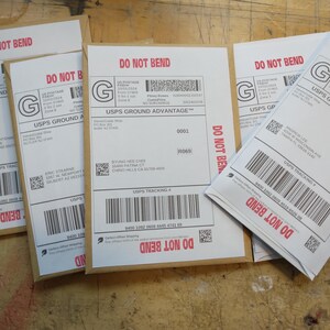 May include: Four white envelopes with brown paper backing. Each envelope has a USPS Ground Advantage label with a tracking number, a red "DO NOT BEND" warning, and a green "G" logo. The text "USPS Ground Advantage" is printed on each label.