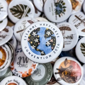 Könnte beinhalten: Ein runder weißer Ansteckbutton mit einer blauen Globus-Illustration, die mit kleinen weißen Blumen verziert ist. Der Text "ETERNAL HAPPINESS CONVENTION 2026" ist in schwarzen Großbuchstaben um den kreisförmigen Rand des Designs gedruckt.