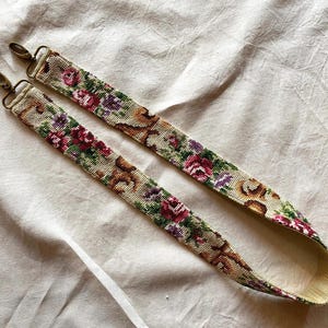 Könnte beinhalten: Ein floraler Gobelin-Taschenriemen mit messingfarbenen Verschlüssen. Der Riemen hat einen beigefarbenen Hintergrund mit einem sich wiederholenden Muster aus roten, lila und grünen Blumen. Der Riemen ist etwa 102 cm lang.