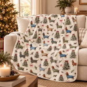 May include: A cream-colored throw blanket featuring a repeating pattern of dachshund dogs wearing sweaters and hats, interspersed with green Christmas trees and red bows. The dogs are depicted in various colors including brown and black.