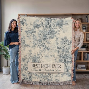 May include: A woven throw blanket with a floral pattern in shades of blue and white. The blanket has a fringe border and a personalized label that reads "Best Mom Ever" with the names "Mom", "Natalie", and "Lola".
