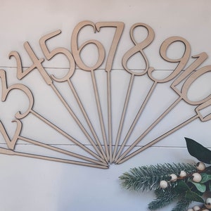 May include: Wooden numbers 1 to 10 on sticks, arranged in a fan shape on a white surface. The numbers are in a cursive font and are a light brown colour.