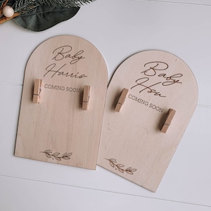 Puede incluir: Dos letreros de madera con el texto "Baby Harris Coming Soon" y "Baby Hsu Coming Soon" con hojas decorativas en la parte inferior. Cada letrero tiene dos pinzas de madera.