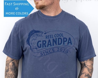 May include: A blue t-shirt with a graphic of a fish jumping out of the water. The text "Reel Cool Grandpa Since 2020" is printed on the shirt.