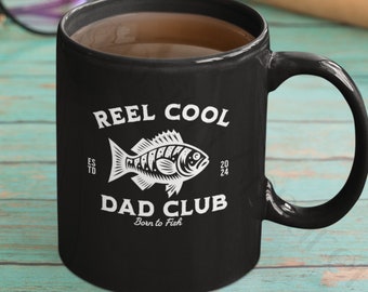 May include: Black ceramic coffee mug with a white design that says "Reel Cool Dad Club Born to Fish" and features a stylised fish illustration.