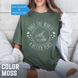 May include: A green t-shirt with the text "Bake the World a Better Place" in white. The design includes a mixing bowl, whisk, and eggs.