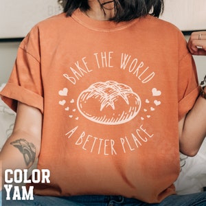 May include: A burnt orange t-shirt with a white graphic of a loaf of bread and the text "Bake the world a better place".