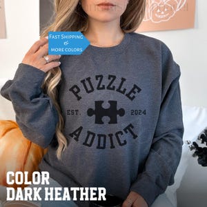 May include: Dark heather grey sweatshirt with a black graphic that says "Puzzle Addict" with a puzzle piece in the center and the text "Est. 2024".