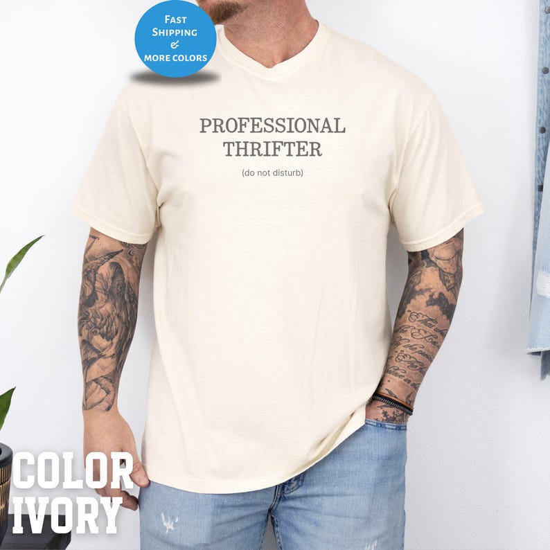 May include: Ivory-colored t-shirt with the text "PROFESSIONAL THRIFTER" and the phrase "(do not disturb)" printed in gray. The shirt has a crew neck and short sleeves. A blue circle with white text that says "FAST SHIPPING & MORE COLORS" is in the upper left corner.